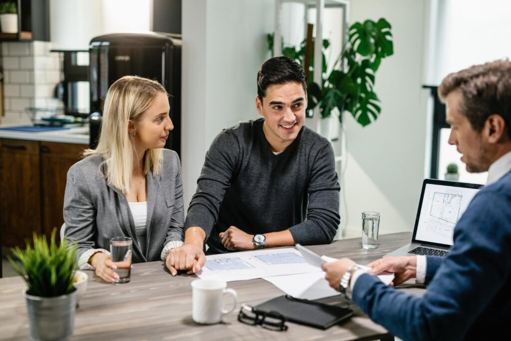 A Young Couple Sitting Across A Desk From A Professional Advisor In A Modern Office, Reviewing Documents And Smiling During A Consultation
